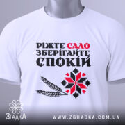 Футболка сало і спокій, біла футболка складена на поверхні, фон світло-сірий, принт з текстом "Ріжте сало зберігайте спокій" у червоному та чорному кольорах, з додатковими графічними елементами, на футболці є етикетка.