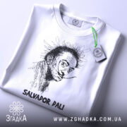 Футболка Сальвадор Далі білого кольору, зображена зверху, на ній підвішений ярлик з зеленим шнурком, фон світло-сірий, принт з портретом Сальвадора Далі в чорному кольорі, підпис 'SALVADOR DALI' внизу.