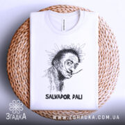 Футболка Сальвадор Далі білого кольору, зображена зверху на плетеній кошику, фон світло-сірий, принт з портретом Сальвадора Далі в чорному кольорі, підпис 'SALVADOR DALI' внизу.