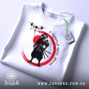 Футболка самурай без дрона, біла, складена на поверхні, фон світлий, принт з чорним самураєм на червоному колі, з зеленим ярликом.