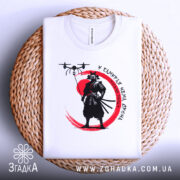 Футболка самурай без дрона, біла, складена на плетеній кошику, фон нейтральний, принт з чорним самураєм на червоному колі та дронами над ним.