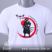 Футболка самурай без дрона, біла, складена на поверхні, фон світлий, принт з чорним самураєм на червоному колі, з зеленим ярликом.