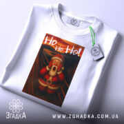 Футболка Санта білого кольору, складена на столі, фон світло-сірий, принт з Сантою в червоному костюмі, який вигукує 'Ho Ho Ho!', з етикеткою на зеленому шнурку.