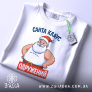 Футболка Санта Клаус одружений, біла, зображена зверху, складена на поверхні, фон світло-сірий, принт з Сантою в червоній шапці та написом 'ОДРУЖЕНИЙ' на червоному фоні, з етикеткою на зеленій стрічці.