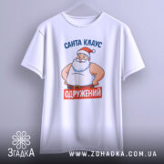 Футболка Санта Клаус одружений, біла, підвішена на вішалці, фон світло-сірий, принт з Сантою в червоній шапці та написом 'ОДРУЖЕНИЙ' на червоному фоні.