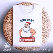 Футболка Санта Клаус одружений, біла, складена на плетеній кошику, фон світло-сірий, принт з Сантою в червоній шапці та написом 'ОДРУЖЕНИЙ' на червоному фоні.