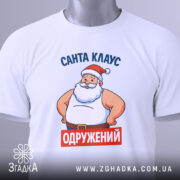 Футболка Санта Клаус одружений, біла, зображена зверху, складена на поверхні, фон світло-сірий, принт з Сантою в червоній шапці та написом 'ОДРУЖЕНИЙ' на червоному фоні, з етикеткою на зеленій стрічці.
