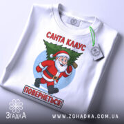 Футболка Санта Клаус повернеться, біла, зображена з верхнього ракурсу, фон світлий, принт з Санта Клаусом, який біжить з ялинкою, текст червоним кольором: 'Санта КЛАУС ПОВЕРНЕТЬСЯ!', на футболці є етикетка з зеленим шнурком.