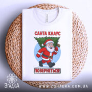 Футболка Санта Клаус повернеться, біла, зображена з верхнього ракурсу, фон нейтральний, принт з Санта Клаусом, який несе ялинку, текст червоним кольором: 'Санта КЛАУС ПОВЕРНЕТЬСЯ!', футболка лежить на плетеній кошику.