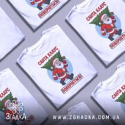 Футболка Санта Клаус повернеться, біла, зображена з фронтального ракурсу, фон нейтральний, принт з веселим Санта Клаусом, який несе ялинку, текст червоним кольором: 'Санта КЛАУС ПОВЕРНЕТЬСЯ!'