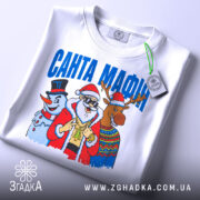 Футболка Санта Мафія білого кольору, складена на поверхні, ракурс зверху, фон світло-сірий, принт з Сантою, сніговиком і оленем, текст "САНТА МАФІЯ" вгорі, на футболці є етикетка.