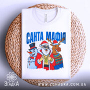 Футболка Санта Мафія білого кольору, складена на плетеній основі, ракурс зверху, фон світло-сірий, принт з Сантою, сніговиком і оленем, текст "САНТА МАФІЯ" вгорі.