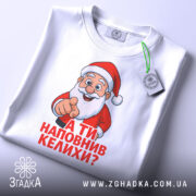 Футболка Санта наповнив келихи, біла футболка, зображена зверху, принт Санти з вказівним жестом, текст червоним кольором: 'А ТИ НАПОВНИВ КЕЛИХИ?', на фоні світло-блакитного кольору, з етикеткою зеленого кольору.