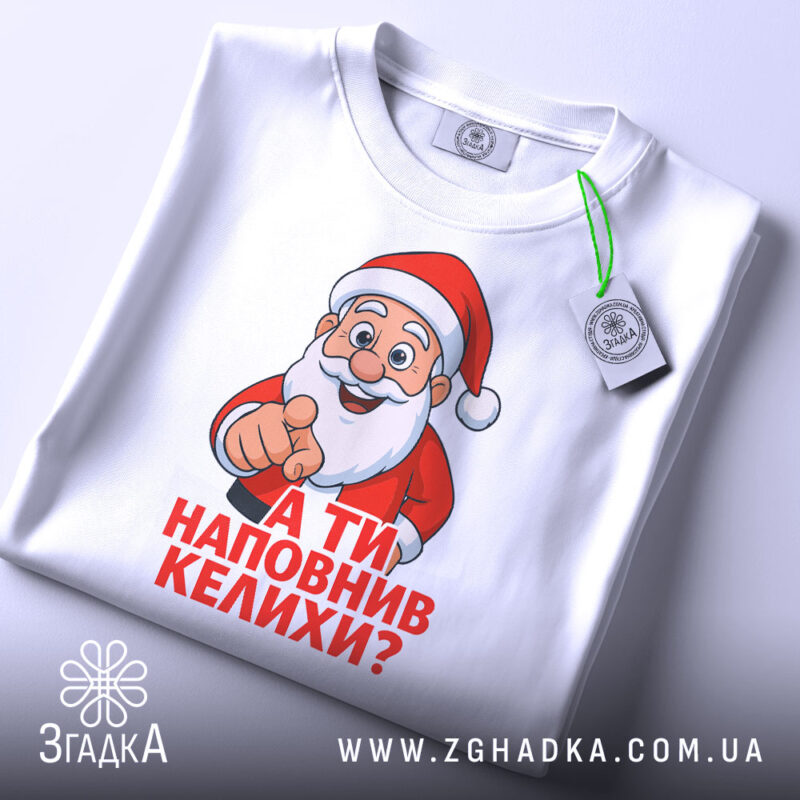 Футболка Санта наповнив келихи, біла футболка, зображена зверху, принт Санти з вказівним жестом, текст червоним кольором: 'А ТИ НАПОВНИВ КЕЛИХИ?', на фоні світло-блакитного кольору, з етикеткою зеленого кольору.