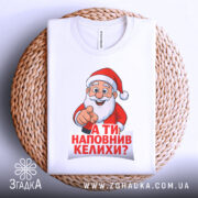 Футболка Санта наповнив келихи, біла футболка, зображена зверху на плетеній кошику, принт Санти з вказівним жестом, текст червоним кольором: 'А ТИ НАПОВНИВ КЕЛИХИ?', на фоні світло-блакитного кольору.