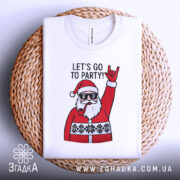 Футболка Санта рок, біла, складена на плетеному кошику, фон світло-сірий, принт з Сантою в окулярах, який піднімає руку в жесті рокера, текст "LET'S GO TO PARTY!" над принтом.