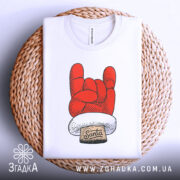 Футболка Санта рок, біла, зображена зверху, на фоні світло-сірого кольору, принт червоного кольору у формі руки з жестом рок, з написом 'Santa' на етикетці, розташована на плетеній основі.