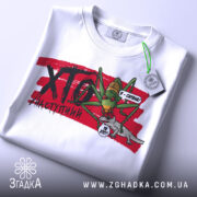 Футболка сарана, біла, зображена на столі, фон світло-сірий, принт з зеленим саранчою та сірим ящіркою, на футболці є етикетка з зеленим шнурком.