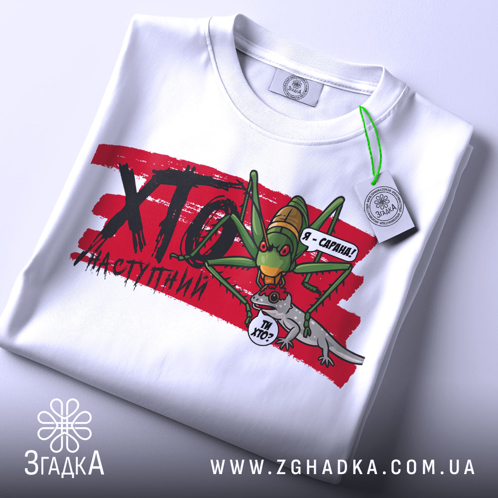 Футболка сарана, біла, зображена на столі, фон світло-сірий, принт з зеленим саранчою та сірим ящіркою, на футболці є етикетка з зеленим шнурком.