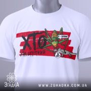 Футболка сарана, біла, зображена на столі, фон світло-сірий, принт з зеленим саранчою та сірим ящіркою, на футболці є етикетка з зеленим шнурком.