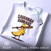 Футболка Savannah Bananas білого кольору, складена на поверхні, фон світло-сірий, принт з жовтою бананою в окулярах та битою, текст 'SAVANNAH BANANAS' над малюнком, на футболці є етикетка з зеленим шнурком.