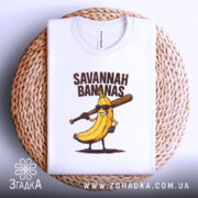 Футболка Savannah Bananas білого кольору, складена на плетеній кошику, фон світло-сірий, принт з жовтою бананою в окулярах та битою, текст 'SAVANNAH BANANAS' над малюнком.