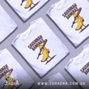 Футболка Savannah Bananas білого кольору, зображена з близького ракурсу на манекені, фон градієнтний сірий, принт з жовтою бананою в окулярах та битою, текст 'SAVANNAH BANANAS' над малюнком.
