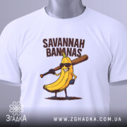 Футболка Savannah Bananas білого кольору, складена на поверхні, фон світло-сірий, принт з жовтою бананою в окулярах та битою, текст 'SAVANNAH BANANAS' над малюнком, на футболці є етикетка з зеленим шнурком.