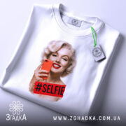 Футболка селфі Мерилін, біла, складена на поверхні, фон світло-сірий, принт з усміхненою Мерилін Монро, яка тримає червоний телефон, з написом #SELFIE на червоному фоні, з етикеткою на комірці.