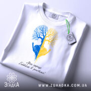 Футболка серце в ритмі, біла, складена на столі, фон світло-сірий, принт з двох дерев, одне синє, інше жовте, з руками, що тримають серце, під написом 'Моє серце б'ється в ритмі!', з етикеткою на зеленій стрічці.