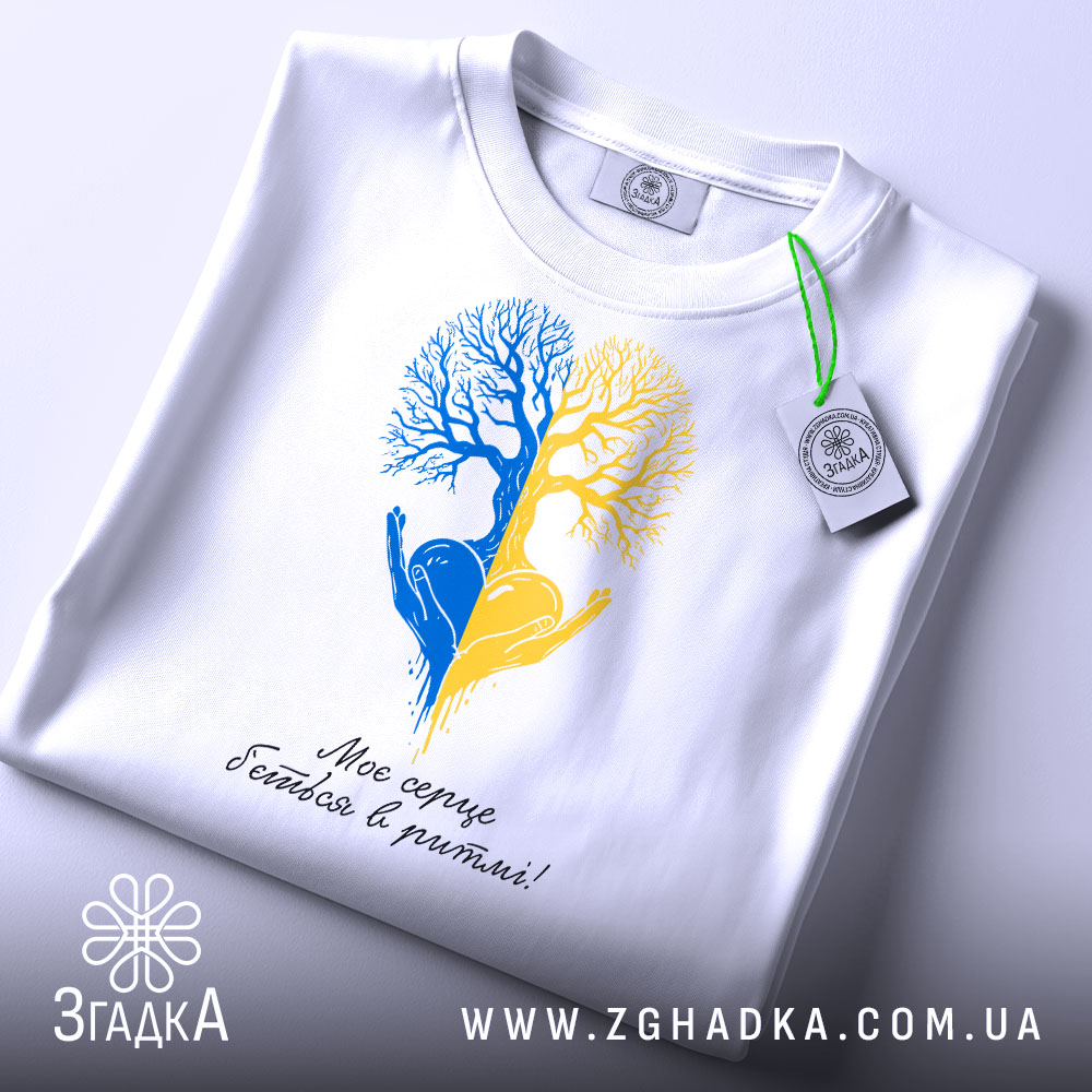 Футболка серце в ритмі, біла, складена на столі, фон світло-сірий, принт з двох дерев, одне синє, інше жовте, з руками, що тримають серце, під написом 'Моє серце б'ється в ритмі!', з етикеткою на зеленій стрічці.