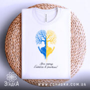 Футболка серце в ритмі, біла, складена на плетеному кошику, фон світлий, принт з двох дерев, одне синє, інше жовте, з руками, що тримають серце, під написом 'Моє серце б'ється в ритмі!'.