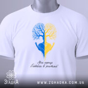 Футболка серце в ритмі, біла, складена на столі, фон світло-сірий, принт з двох дерев, одне синє, інше жовте, з руками, що тримають серце, під написом 'Моє серце б'ється в ритмі!', з етикеткою на зеленій стрічці.