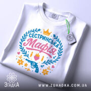 Футболка сестринська мафія білого кольору, складена на поверхні з легким нахилом, фон світло-сірий, принт яскравий з написом 'Сестринська Мафія', пістолетом, кулі, квіти та сердечка, на комірці є етикетка.