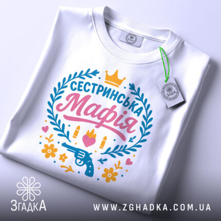 Футболка сестринська мафія білого кольору, складена на поверхні з легким нахилом, фон світло-сірий, принт яскравий з написом 'Сестринська Мафія', пістолетом, кулі, квіти та сердечка, на комірці є етикетка.