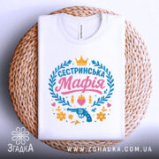 Футболка сестринська мафія білого кольору, складена на круглому плетеному кошику, фон світло-сірий, принт яскравий з написом 'Сестринська Мафія', пістолетом, кулі, квіти та сердечка.