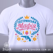Футболка сестринська мафія білого кольору, складена на поверхні з легким нахилом, фон світло-сірий, принт яскравий з написом 'Сестринська Мафія', пістолетом, кулі, квіти та сердечка, на комірці є етикетка.