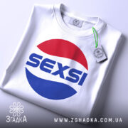 Футболка SEXSI білого кольору, складена на поверхні, з великим принтом у вигляді червоного та синього кола, на якому написано 'SEXSI' великими літерами, на фоні світло-сірого кольору, з видимою етикеткою з зеленим шнурком.