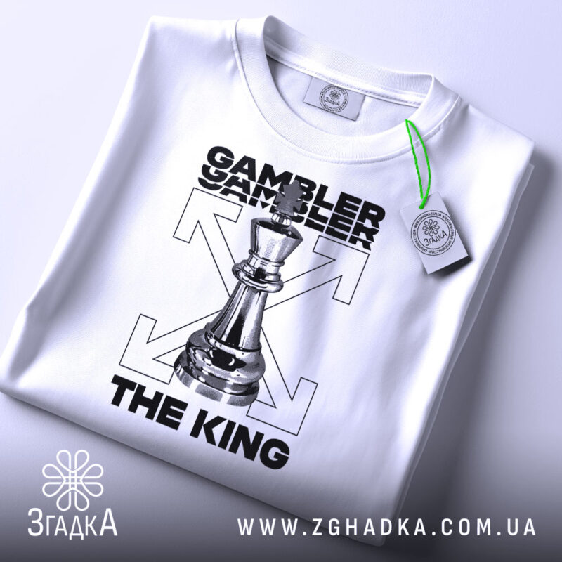 Футболка шаховий король, біла, зображена в горизонтальному положенні, фон світло-сірий, принт з шаховою фігурою короля та текстом 'GAMBLER THE KING', на футболці є етикетка з зеленим шнурком.