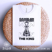 Футболка шаховий король, біла, зображена в горизонтальному положенні на плетеній основі, фон світло-сірий, принт з шаховою фігурою короля та текстом 'GAMBLER THE KING'.