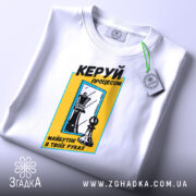 Футболка шахи керуй процесом, біла, складена на поверхні, фон світлий, принт з шаховими фігурами та текстом у жовтій рамці, з етикеткою.