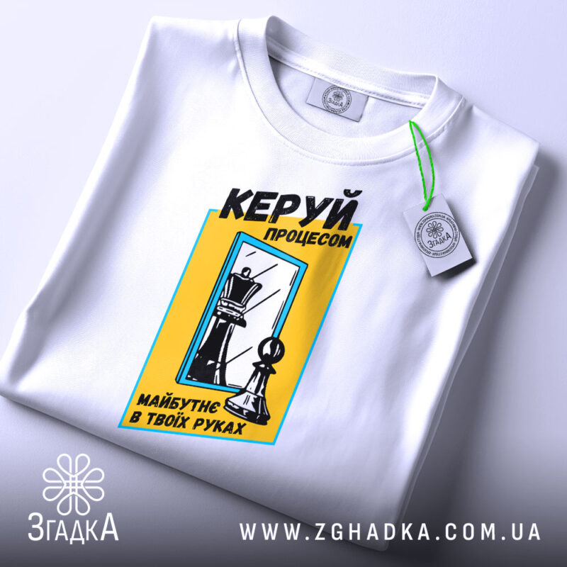 Футболка шахи керуй процесом, біла, складена на поверхні, фон світлий, принт з шаховими фігурами та текстом у жовтій рамці, з етикеткою.