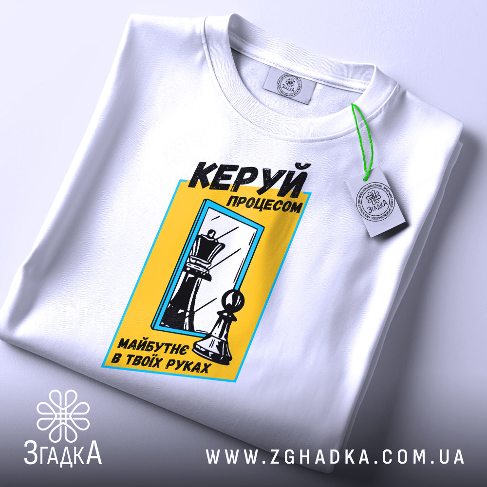 Футболка шахи керуй процесом, біла, складена на поверхні, фон світлий, принт з шаховими фігурами та текстом у жовтій рамці, з етикеткою.