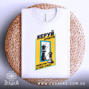 Футболка шахи керуй процесом, біла, складена на плетеній кошику, фон світлий, принт з шаховими фігурами та текстом у жовтій рамці.