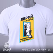 Футболка шахи керуй процесом, біла, складена на поверхні, фон світлий, принт з шаховими фігурами та текстом у жовтій рамці, з етикеткою.