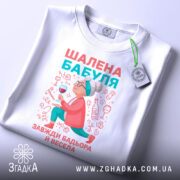 Футболка шалена бабуля, білого кольору, зображена зверху, на ній прикріплена етикетка, фон світло-сірий, принт з бабусею, що тримає келих, в яскравих кольорах.