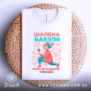 Футболка шалена бабуля, білого кольору, складена на плетеній основі, фон світло-сірий, принт з бабусею, що тримає келих, в яскравих кольорах.