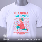 Футболка шалена бабуля, білого кольору, зображена зверху, на ній прикріплена етикетка, фон світло-сірий, принт з бабусею, що тримає келих, в яскравих кольорах.