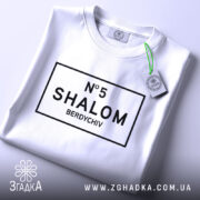 Футболка шалом Бердичів, біла, зображена зверху, на ній видно етикетку з зеленим шнурком, принт чорного кольору з написом 'N°5 SHALOM BERDYCHIV' у прямокутній рамці.
