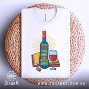 Футболка щасливі люди, білого кольору, зображена зверху на плетеній кошику, фон білий, принт з пляшкою вина, келихом та книгами, яскраві кольори, текст на українській мові.